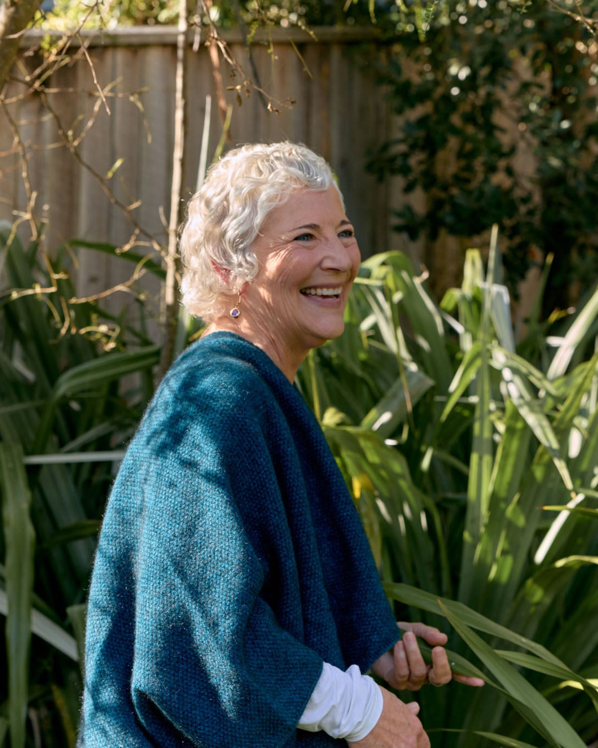 An older woman with short gray hair stands outdoors in a garden, smiling and looking to the side. She is wearing a textured teal blue shawl over a white long-sleeve top.