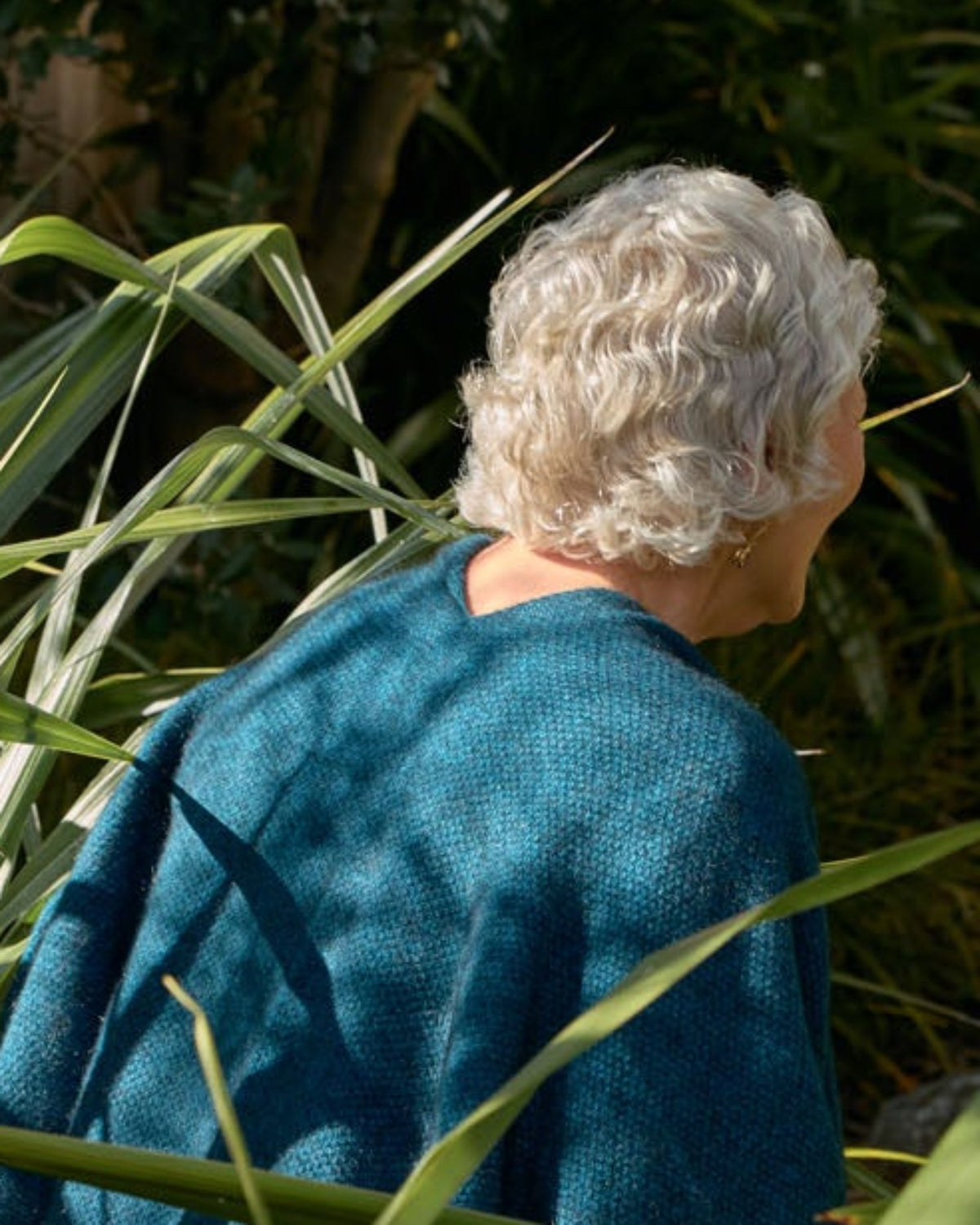 An older person with gray hair seen from behind, wearing a teal blue long-sleeve top and walking through dense green garden foliage.
