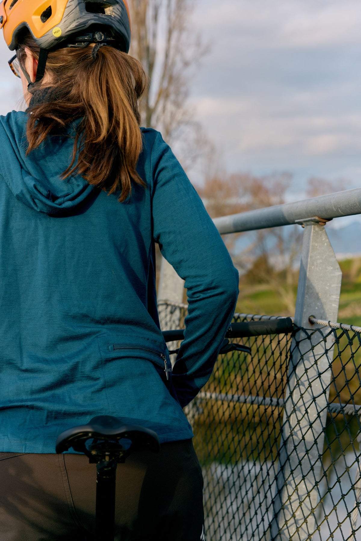 A merino woman wearing a teal long-sleeve athletic hoodie and a black helmet with orange accents, seen from behind as she looks out over a scenic outdoor area with a chain-link fence and trees.