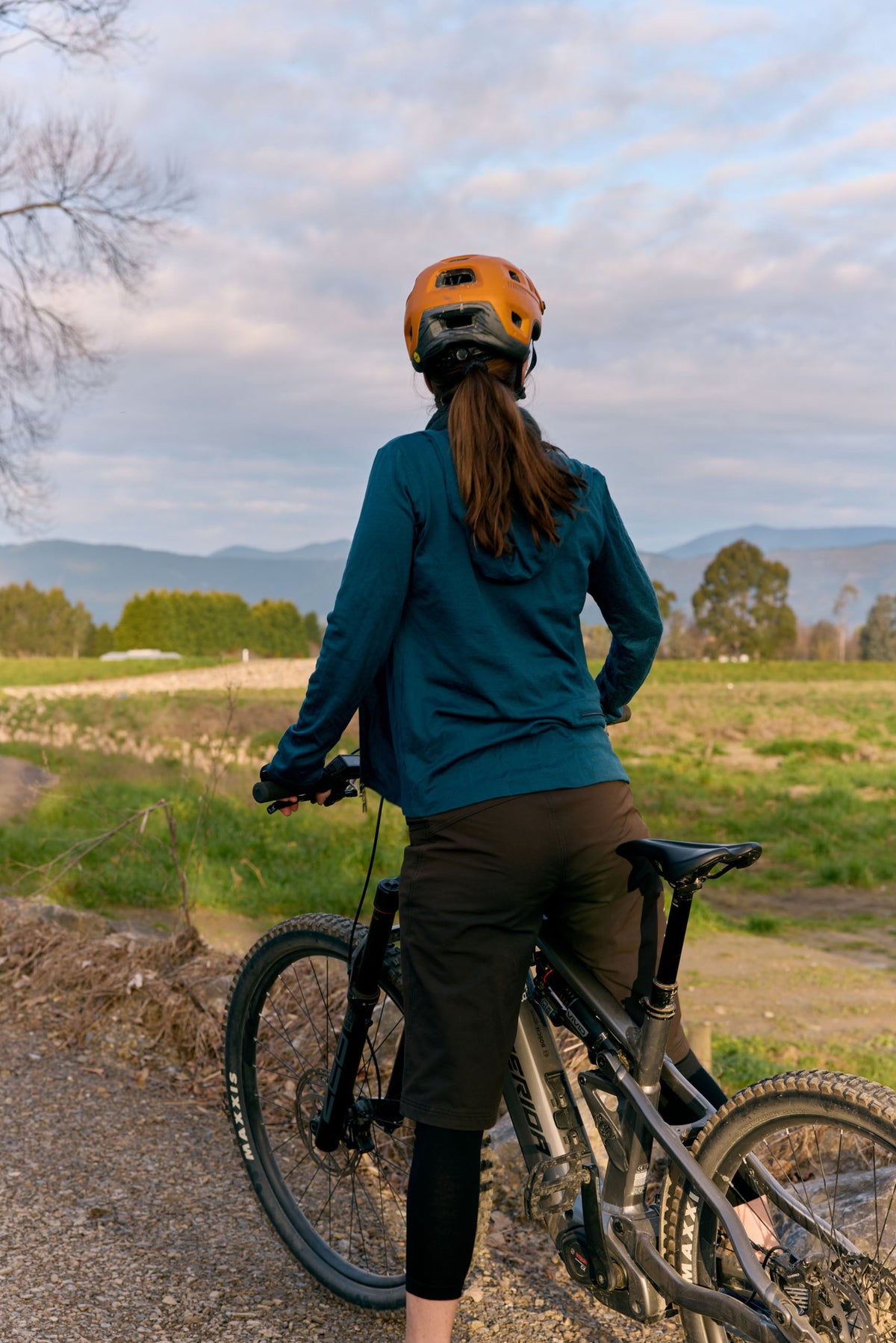 A woman wearing an orange helmet and a merino  teal long-sleeve athletic jacket stands on a gravel path next to a mountain bike, looking out over a grassy field and distant mountains under a cloudy sky.