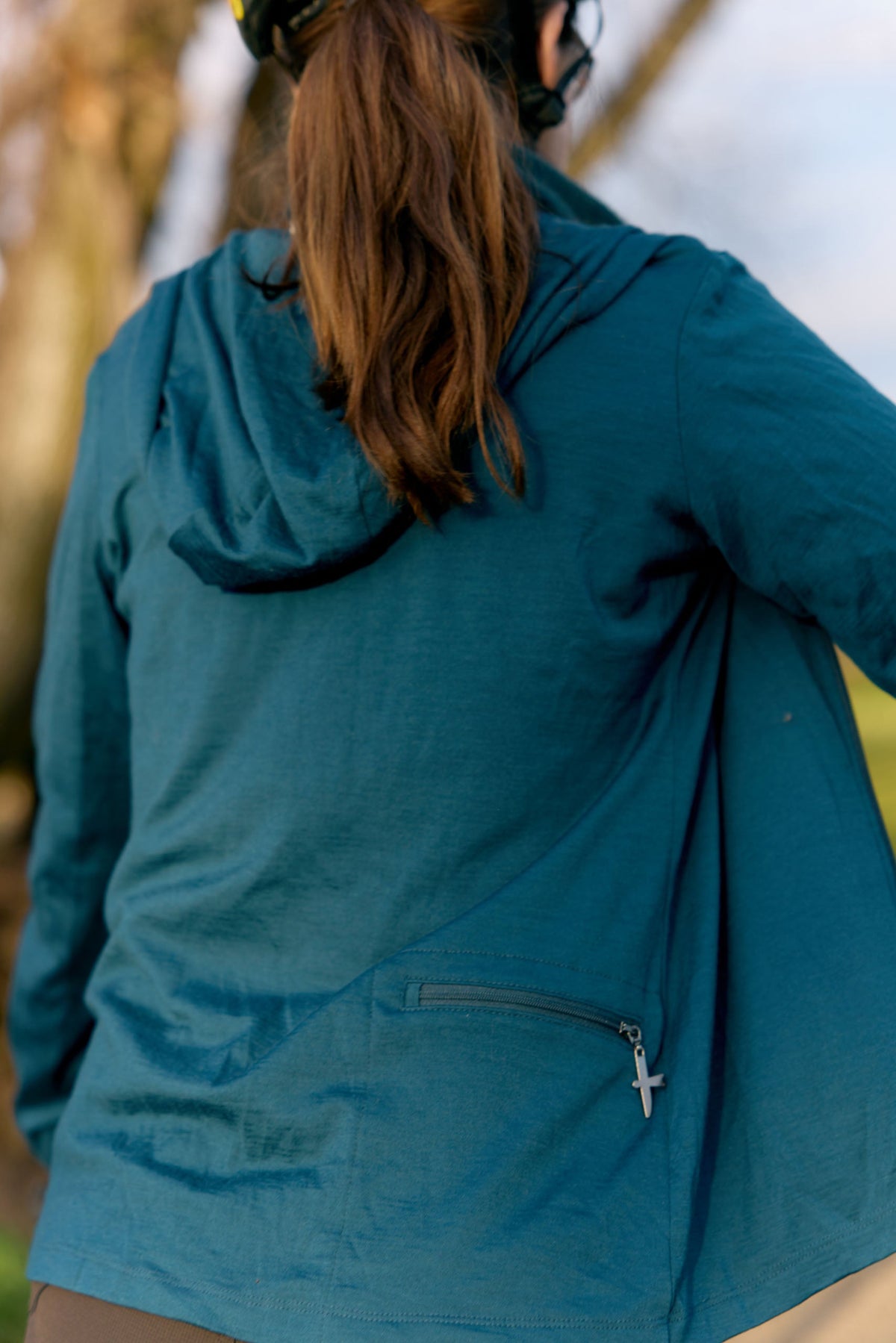 A merino person viewed from behind wearing a teal blue hooded jacket outdoors, with a zippered pocket on the back and brown hair tied up.