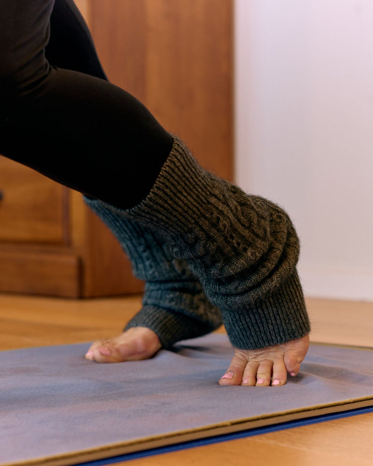 A close-up of a person's feet and lower legs wearing dark green cable-knit leg warmers over black leggings, standing on a gray yoga mat in a room with wooden furniture.