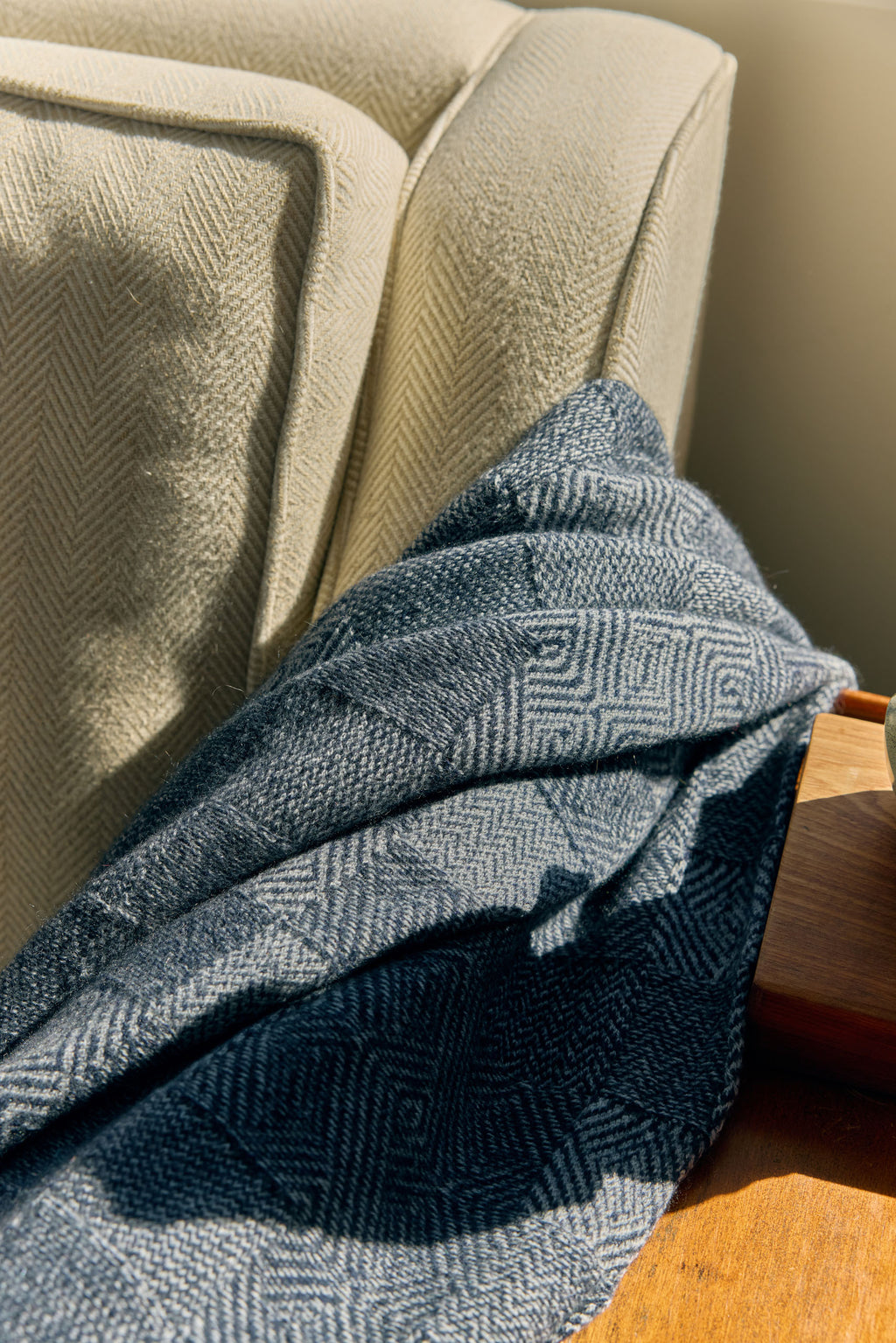 A close-up of a dark blue and gray herringbone patterned throw blanket draped over the arm of a beige armchair in warm natural sunlight.