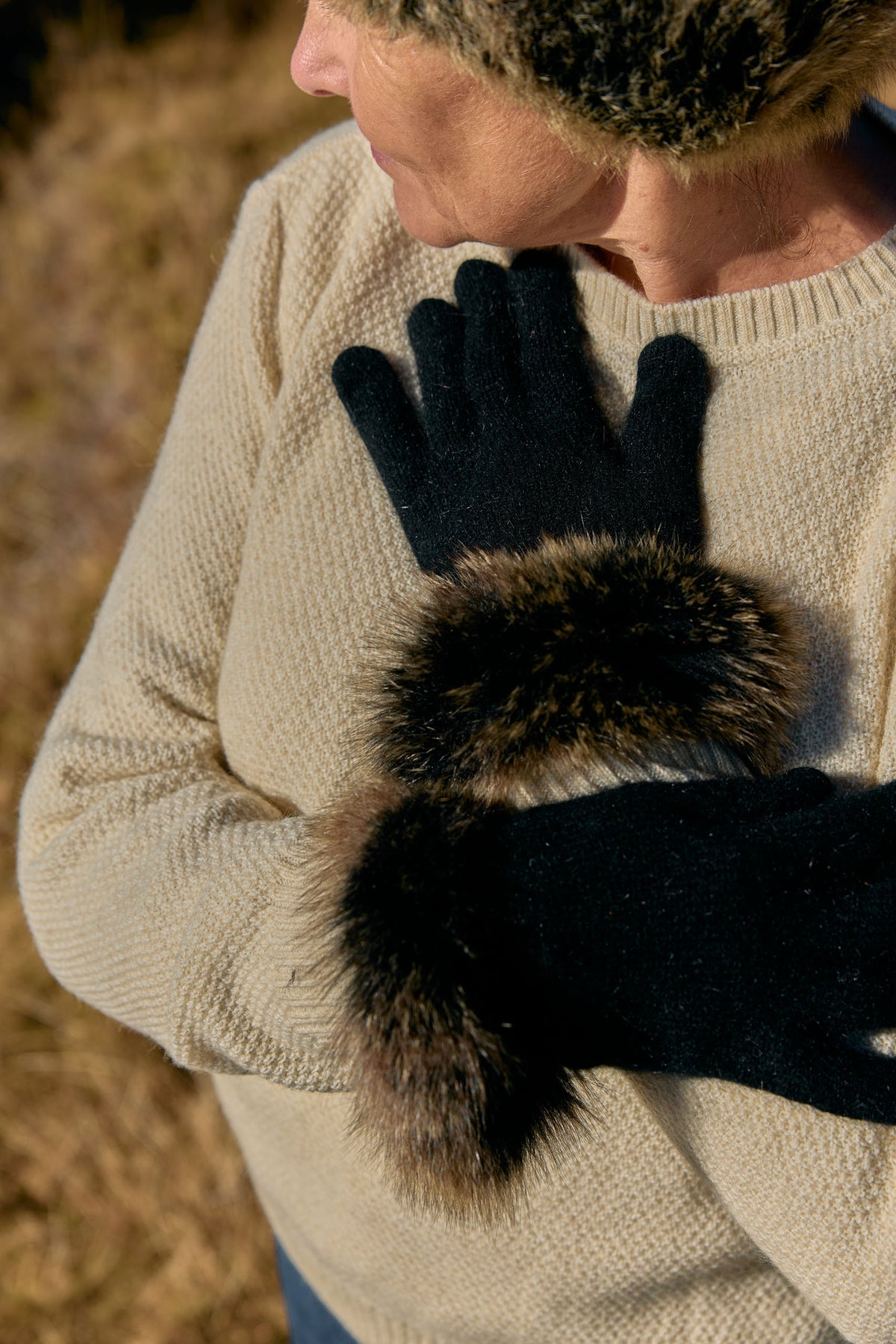 A close-up of a person wearing black knit gloves with dark brown and black fur trim, paired with a cream knit sweater in an outdoor setting.