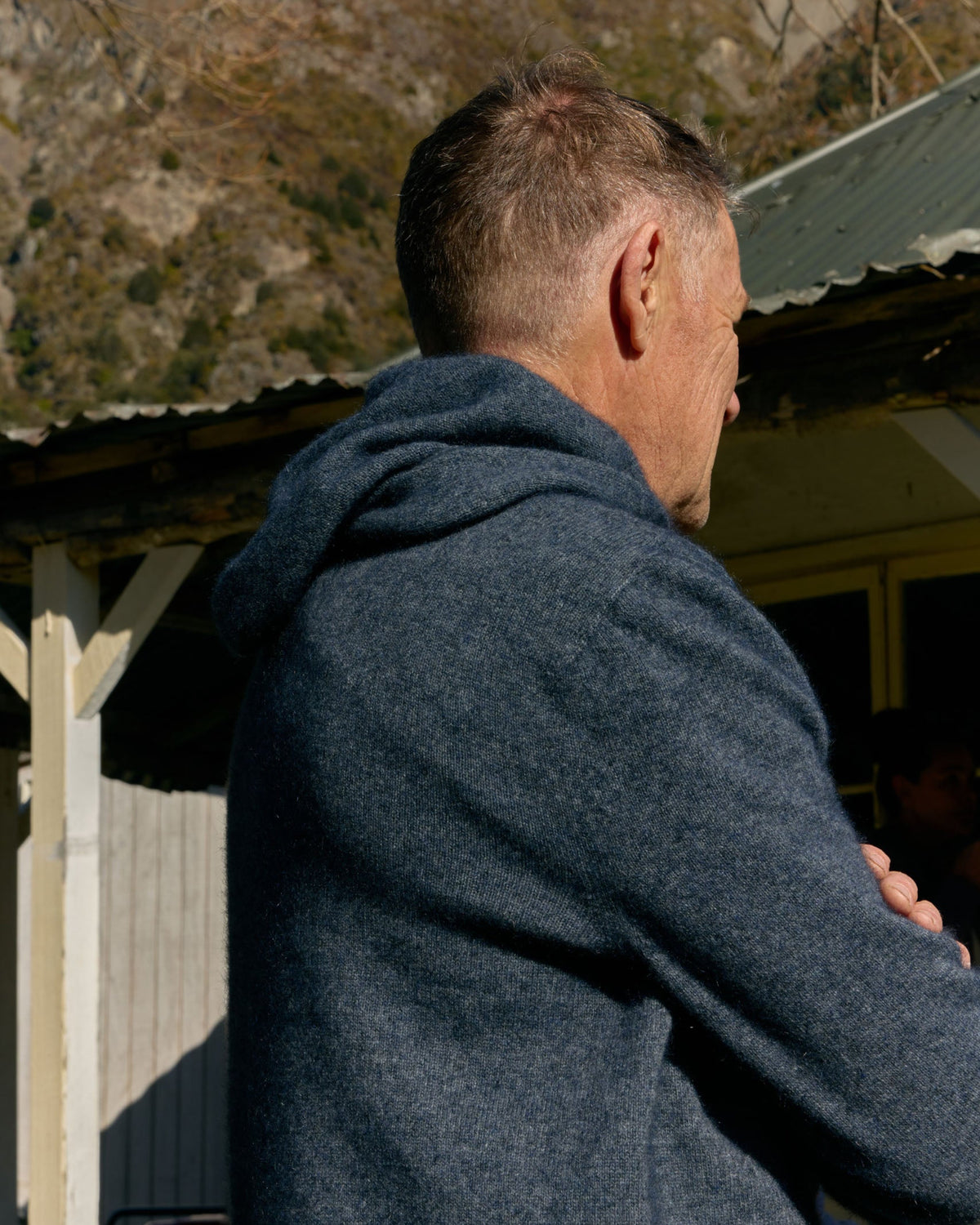 A man viewed from behind wearing a dark heathered blue-gray hooded sweatshirt, standing outdoors in front of a rustic building with a corrugated metal roof and a rocky hillside in the background.