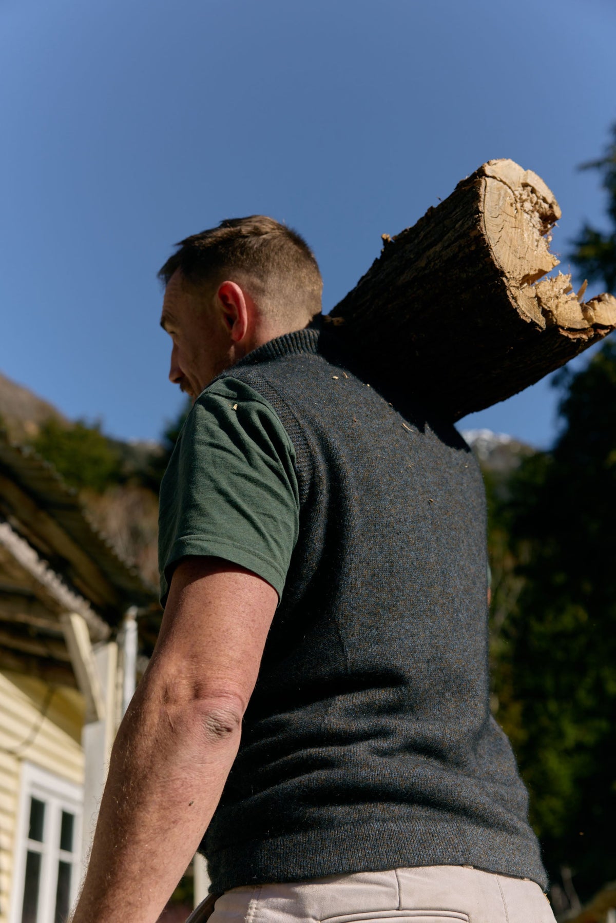 possum merino A man viewed from behind carrying a large cut log on his shoulder, wearing a dark gray knit sweater vest over a green t-shirt and light beige trousers, outdoors under a clear blue sky.