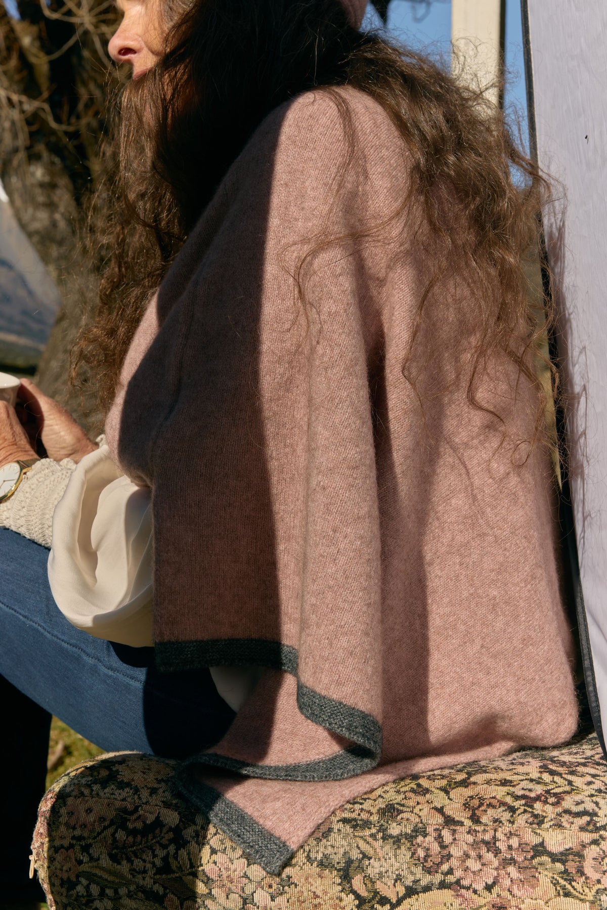 A person outdoors wearing a dusty pink knit poncho or cape with dark gray trim, paired with a cream top and patterned pants.