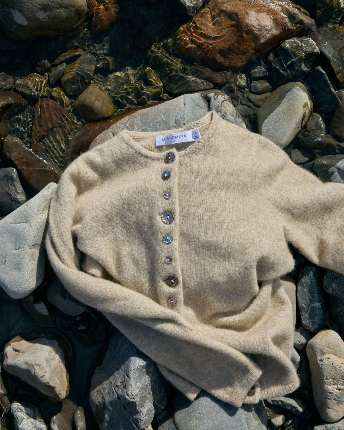 A beige knit cardigan sweater with a buttoned henley neckline, laid flat on a bed of wet river rocks and pebbles.