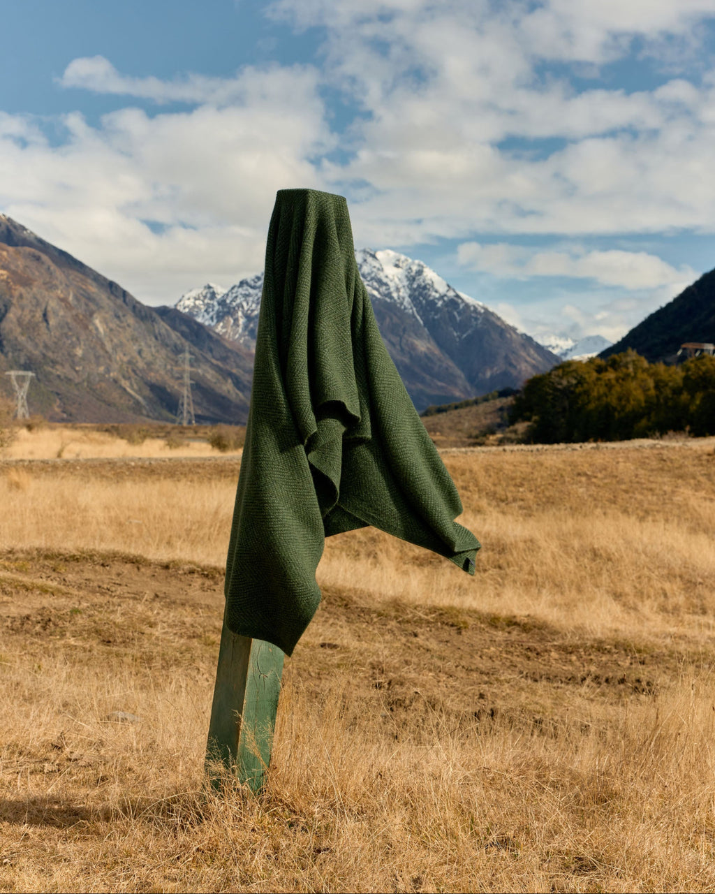 A dark green fleece jacket is draped over a metal post in an open grassy field, with snow-capped mountains and a blue sky with clouds in the background.