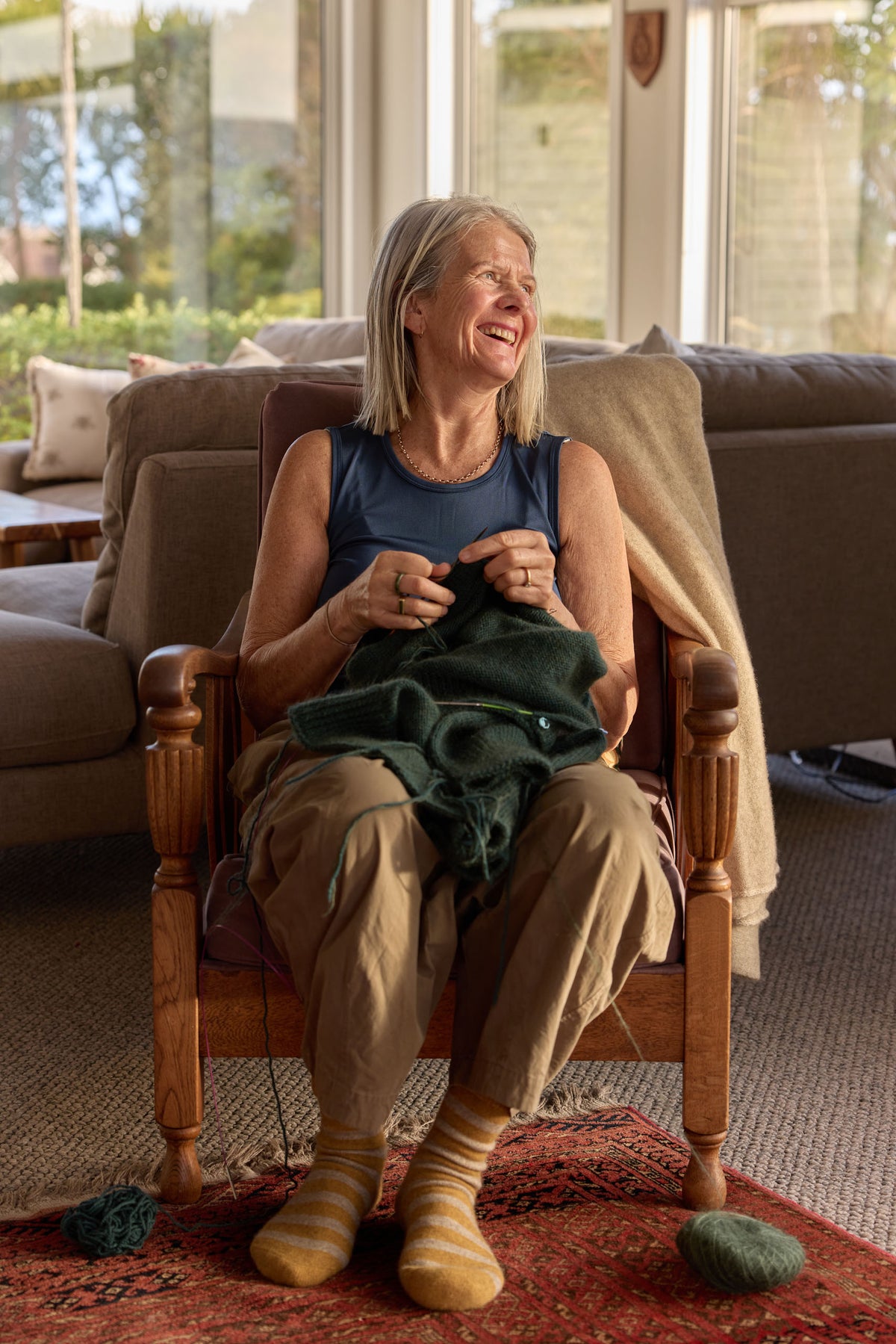 A smiling woman sits in a wooden armchair in a sunlit living room, knitting with a dark green yarn. She wears a blue sleeveless top, tan pants, and striped socks, with a beige throw blanket draped over the chair.