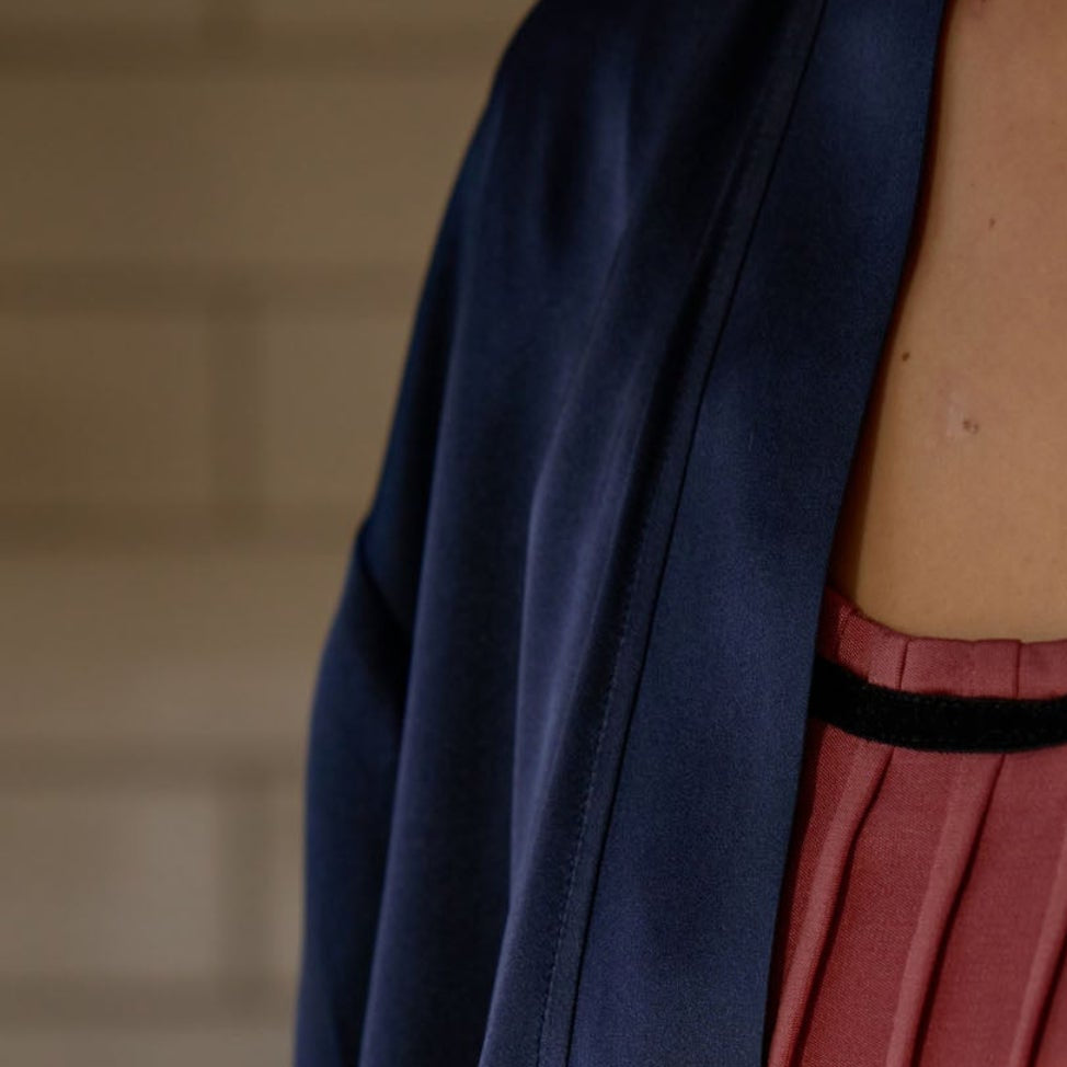 A close-up of a person wearing a navy blue satin-like robe or kimono over a pink pleated top with black trim, standing indoors near a countertop.