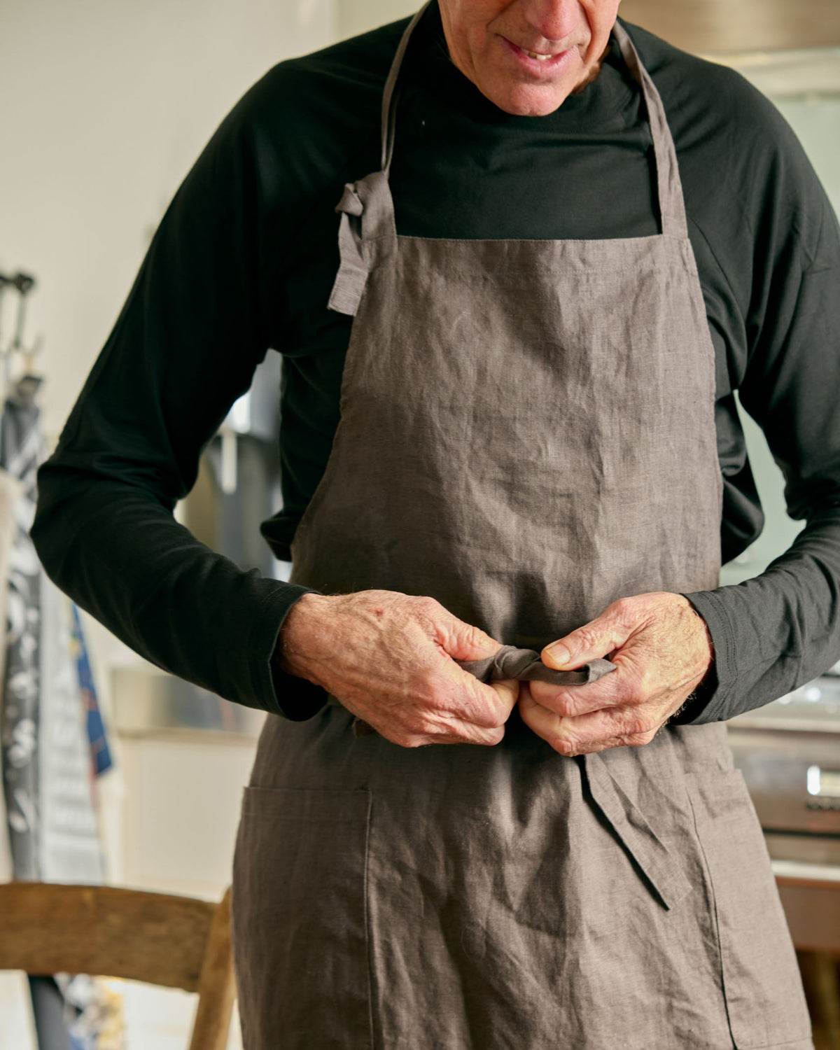 A man wearing a dark gray linen apron over a black long-sleeve shirt, tying the apron at his waist in a kitchen setting.