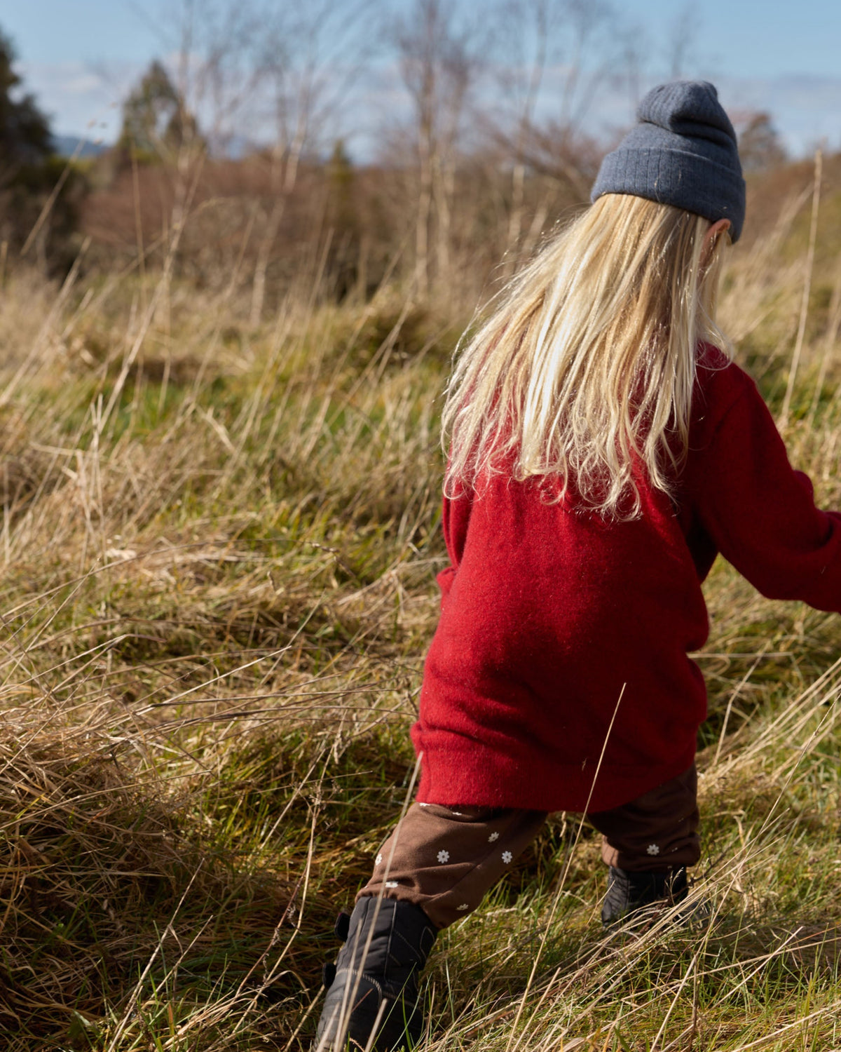 A child seen from behind wearing a red knit sweater, brown pants, black boots, and a dark blue beanie while walking through tall dry grass outdoors.