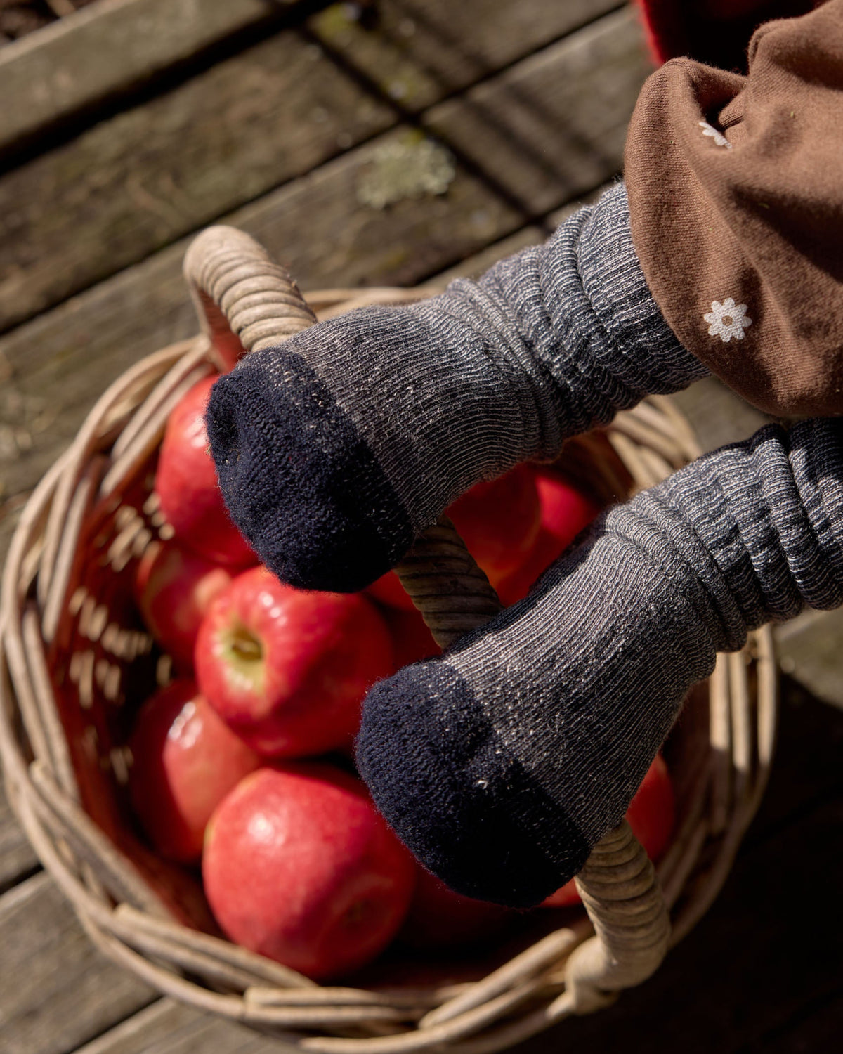 A close-up lifestyle shot of a child's legs wearing gray knit socks with dark navy toe and heel caps, sitting in a wicker basket filled with red apples on a wooden deck.