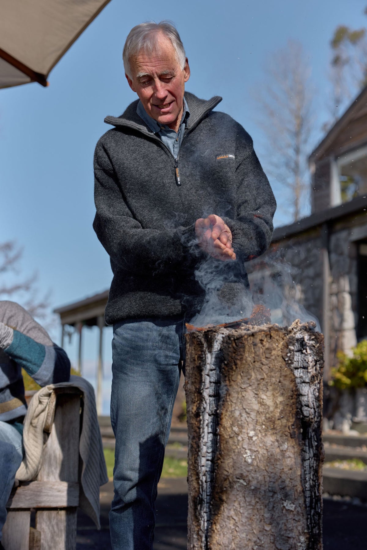 A man wearing a dark gray Possum merino pullover and blue jeans stands outdoors next to a smoking cylindrical container, with a house and trees in the background.