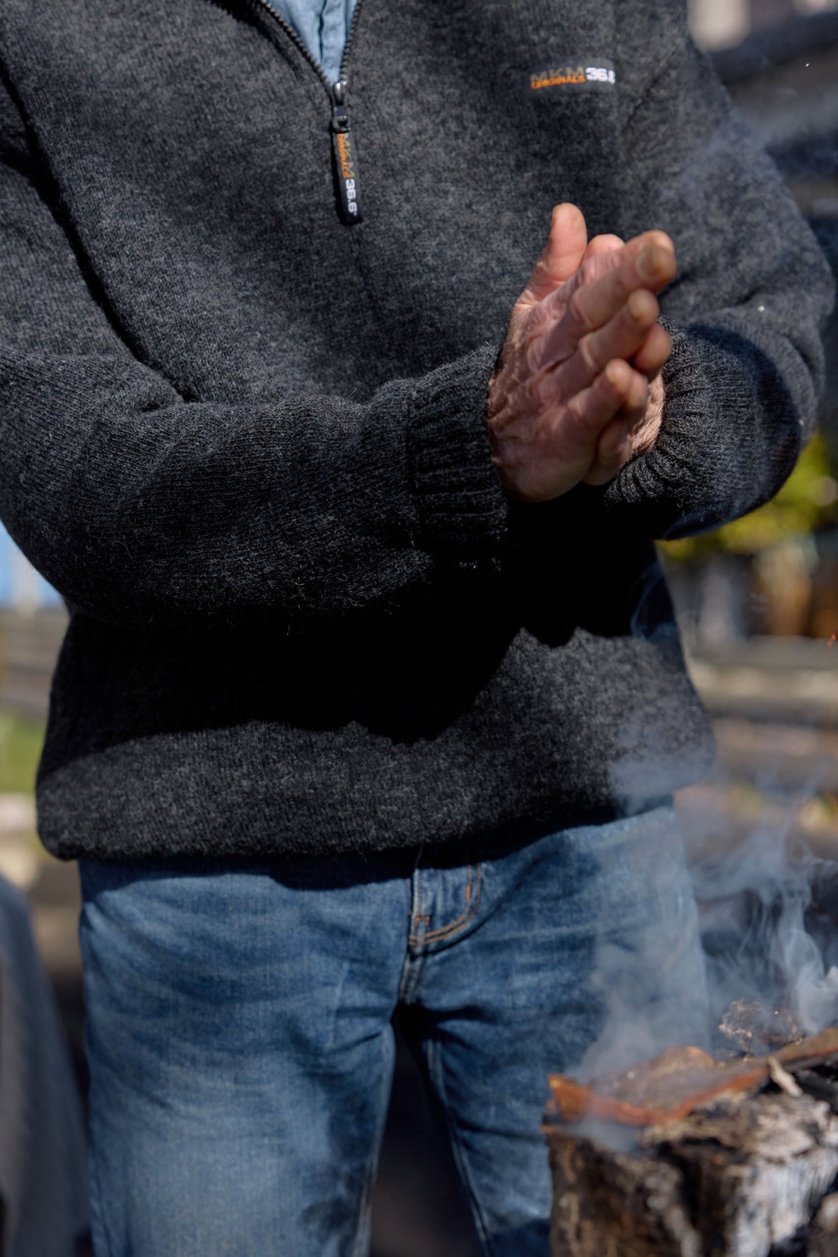 A close-up of a person wearing a dark gray heathered quarter-zip Possum merino  sweater and blue jeans, rubbing their hands together in front of a smoky fire outdoors.