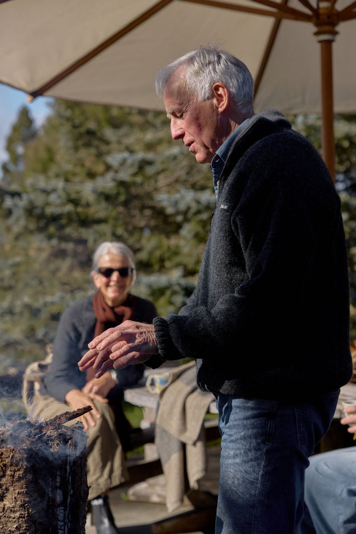 An older man in a dark possum merino  pullover and jeans stands outdoors under a patio umbrella near a grill, with another person seated in the background.