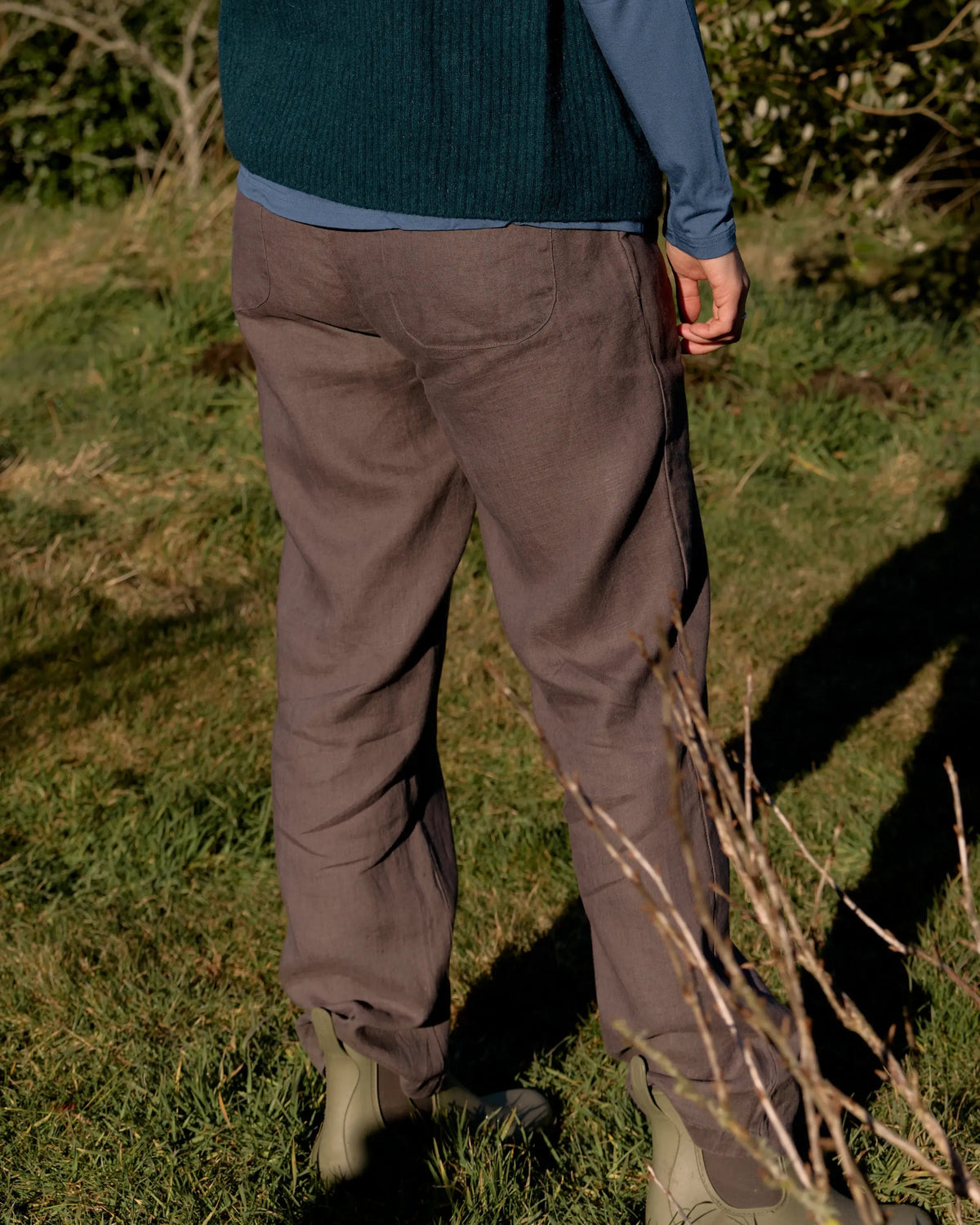A person outdoors on grass wearing loose brown trousers, a dark teal sweater, and a blue long-sleeve shirt, shown from the back with strong sunlight and shadows.