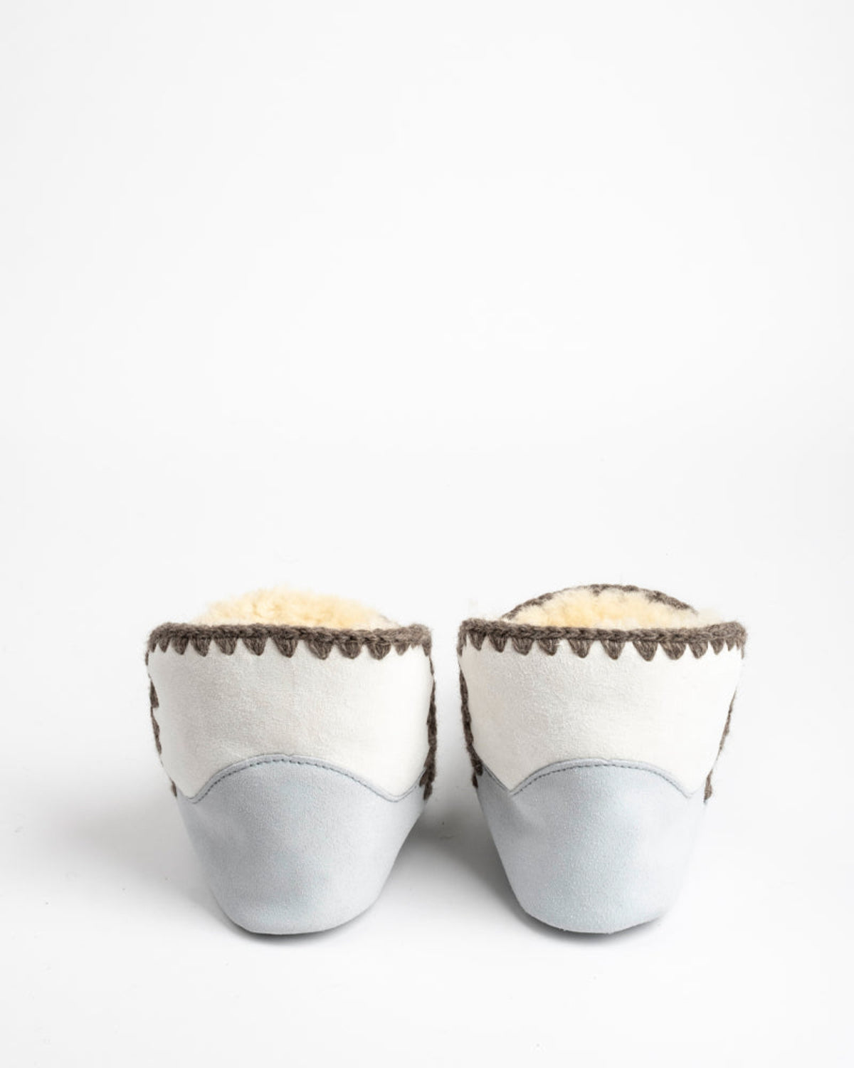 Pair of light gray sheepskin slippers with brown trim on a white background